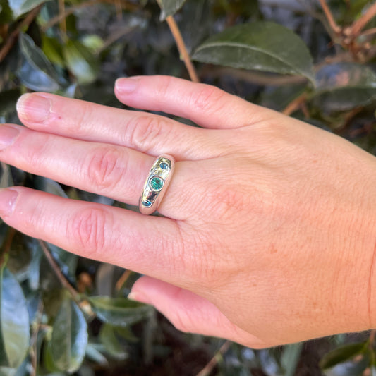 A gold band ring with three small green and blue gemstones, shown worn on a person's hand against a background of green leaves.