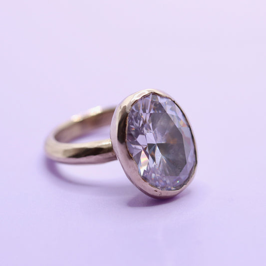 A gold ring with a large, oval-cut clear gemstone in a bezel setting, photographed on a solid light purple background.