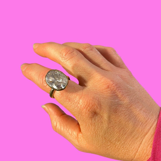 A close-up of a hand wearing a vintage-style ring with a large round clear stone in a gold-toned setting, set against a solid bright pink background.