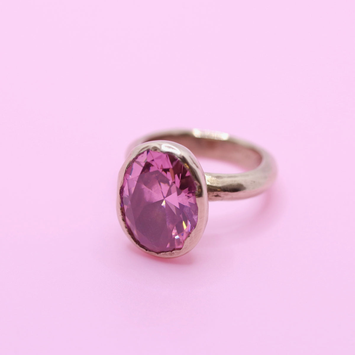A bronze ring featuring a large, oval-cut faceted pink gemstone, set against a solid light pink background.