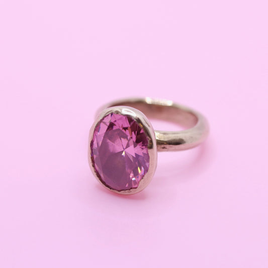 A bronze ring featuring a large, oval-cut faceted pink gemstone, set against a solid light pink background.