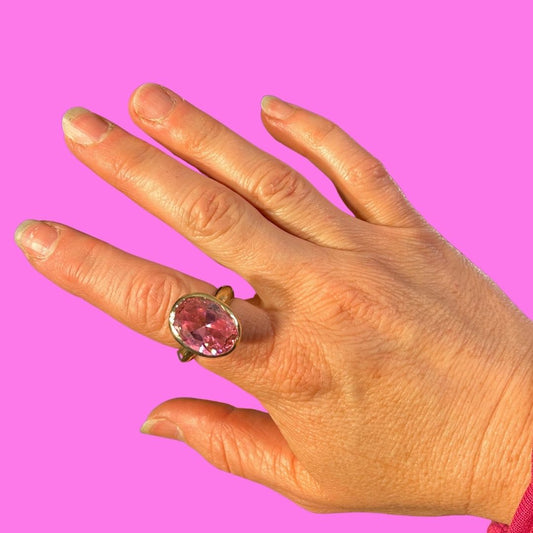 A hand wearing a vintage-style gold ring with a large oval pink gemstone, set against a solid bright pink background.