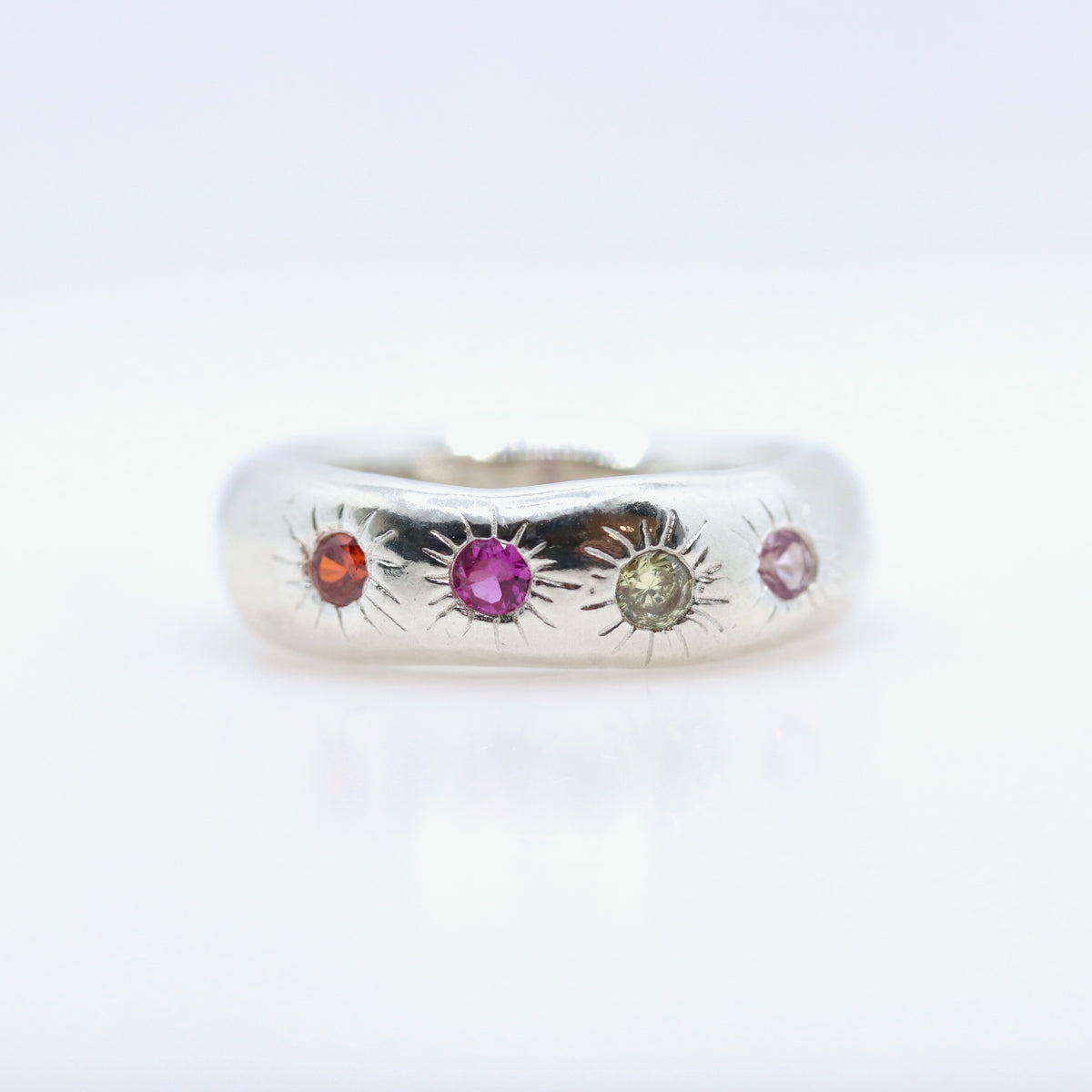 A silver band ring featuring three small sunburst motifs set with colorful gemstones in orange, pink, and green, photographed on a clean white background.