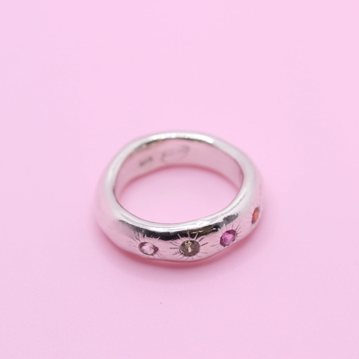 A silver band ring with small colorful gemstones set along the front, photographed on a solid pink background.