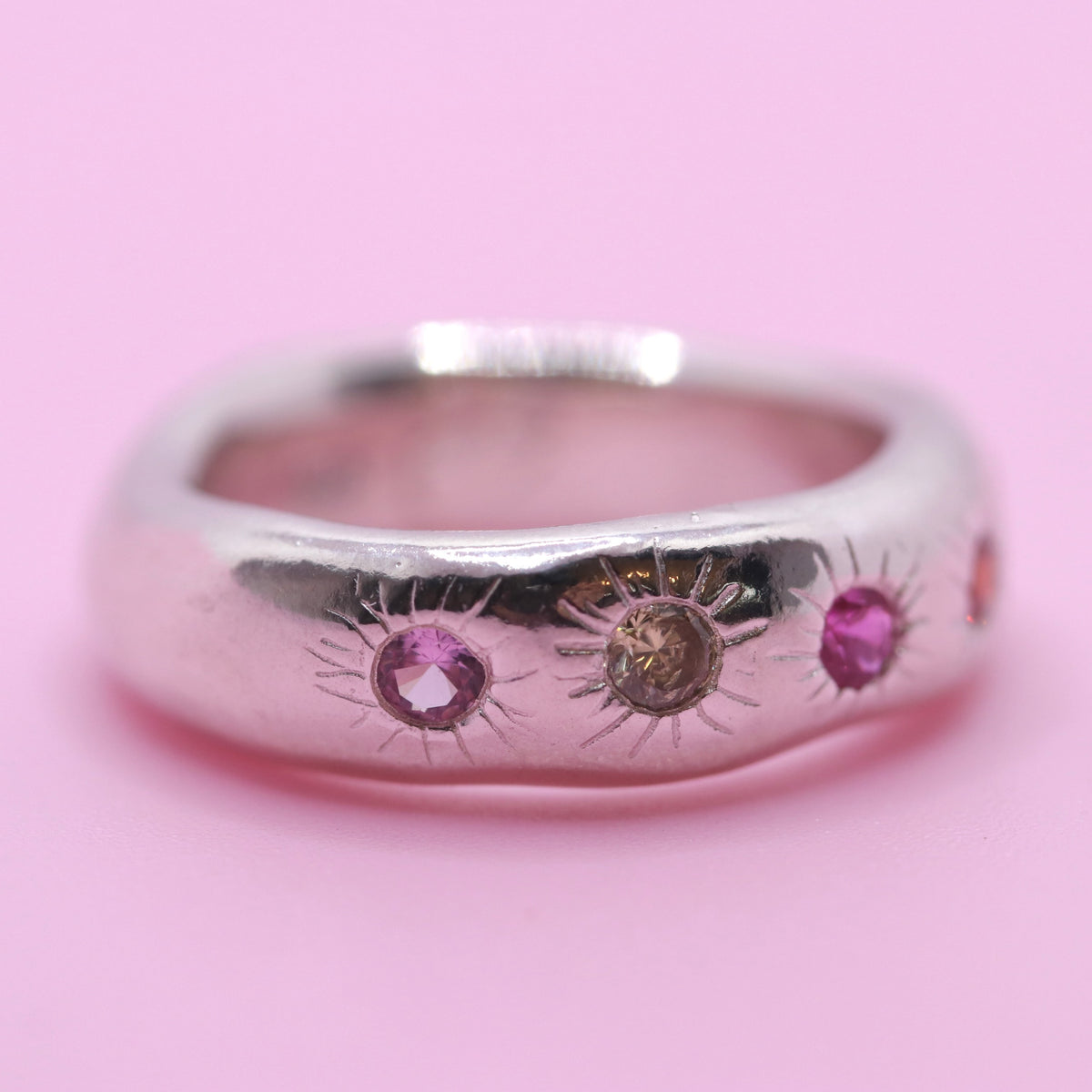 A polished silver ring featuring three sunburst motifs with pink and brown gemstones, set against a solid pink background.