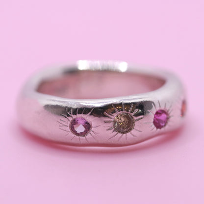 A polished silver ring featuring three sunburst motifs with pink and brown gemstones, set against a solid pink background.