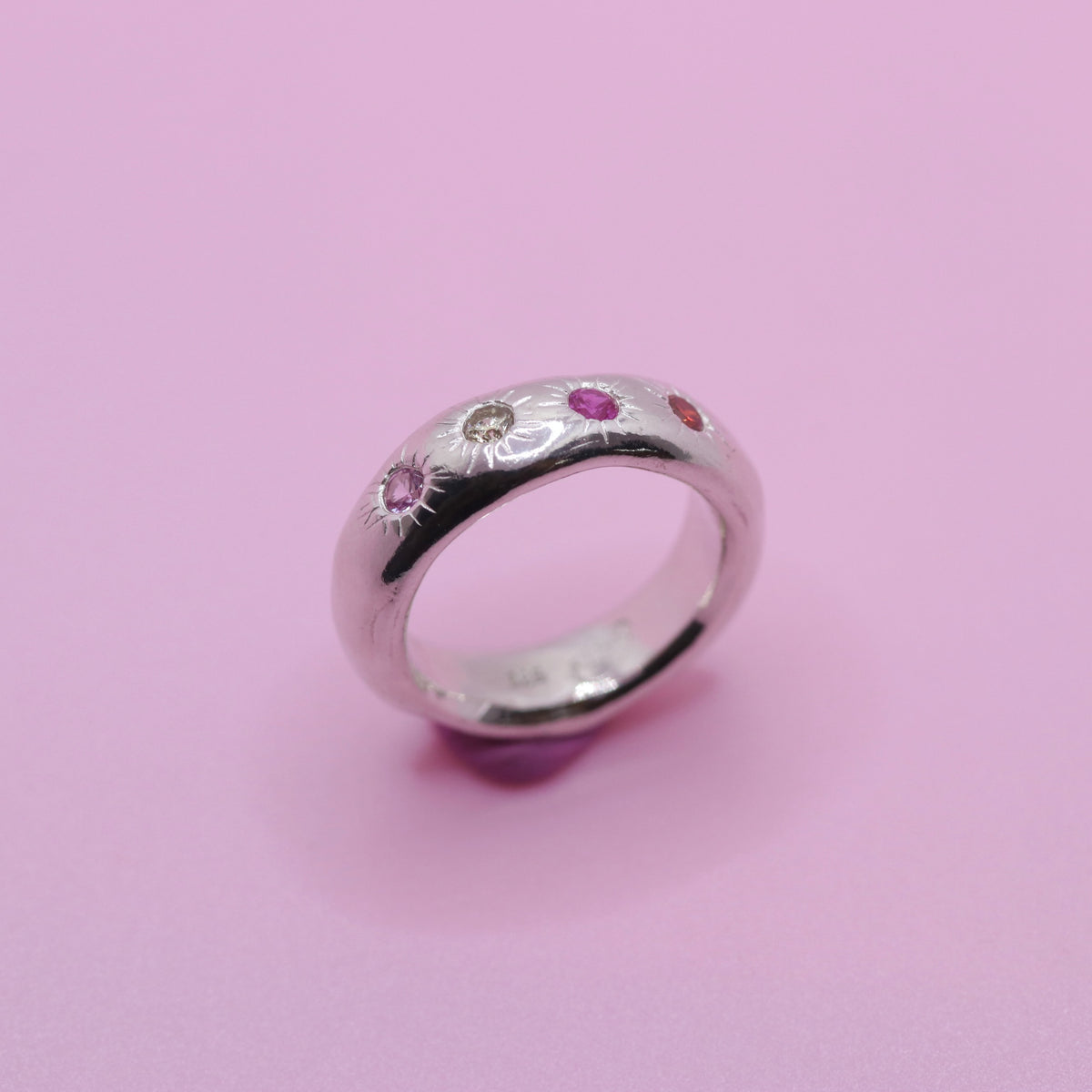 A sterling silver band ring featuring small, colorful gemstone sun motifs in pink, red, and clear, set against a solid pink background.