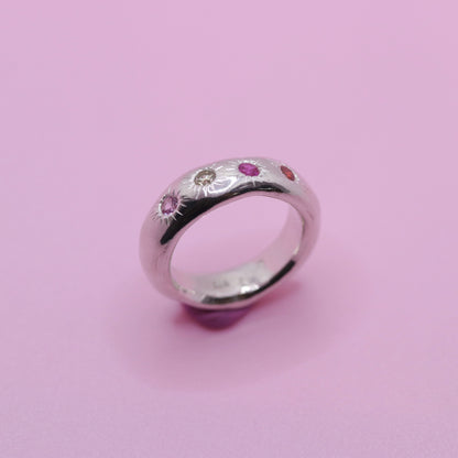 A sterling silver band ring featuring small, colorful gemstone sun motifs in pink, red, and clear, set against a solid pink background.