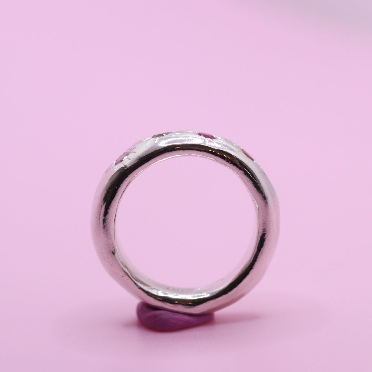 A polished silver ring with a textured, organic shape and small embedded pink gemstones, displayed on a small purple stand against a solid pink background.