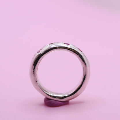 A polished silver ring with a textured, organic shape and small embedded pink gemstones, displayed on a small purple stand against a solid pink background.