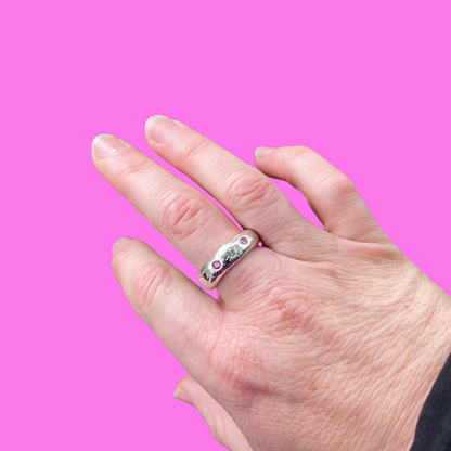 A silver band ring with small pink and green gemstones, worn on a person's middle finger against a solid bright pink background.