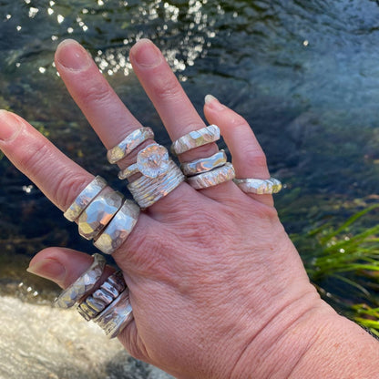 A hand wearing multiple layered silver rings with textured, hammered finishes, photographed outdoors against a blurred background of water and greenery.