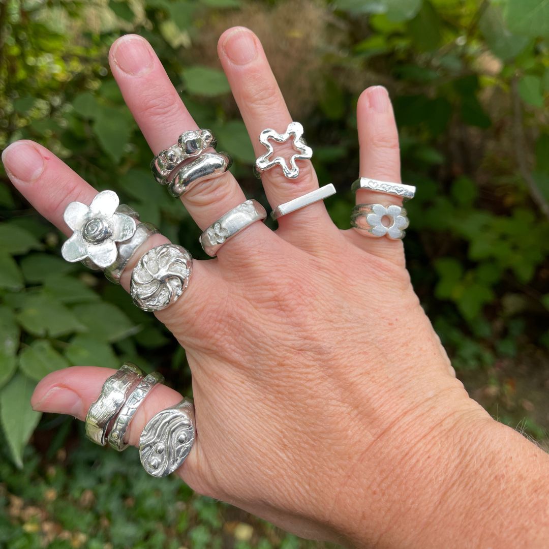 A hand wearing multiple sterling silver rings with various organic and floral designs, photographed outdoors against a blurred green foliage background.