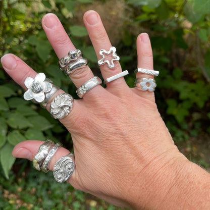 A hand wearing multiple sterling silver rings with various organic and floral designs, photographed outdoors against a blurred green foliage background.