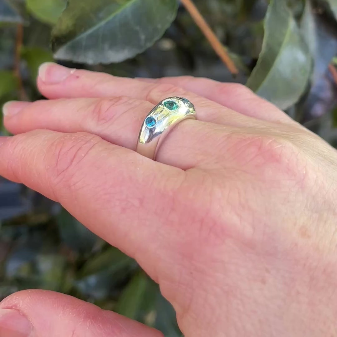 Chunky silver ring with blue and green gems on had with dark green leaves in background.